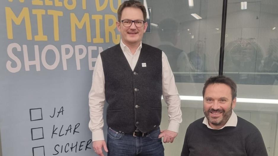 2 Personen, Männer mittleren Alters, eine Person steht, die andere sitzt im Rollstuhl, alle lachen